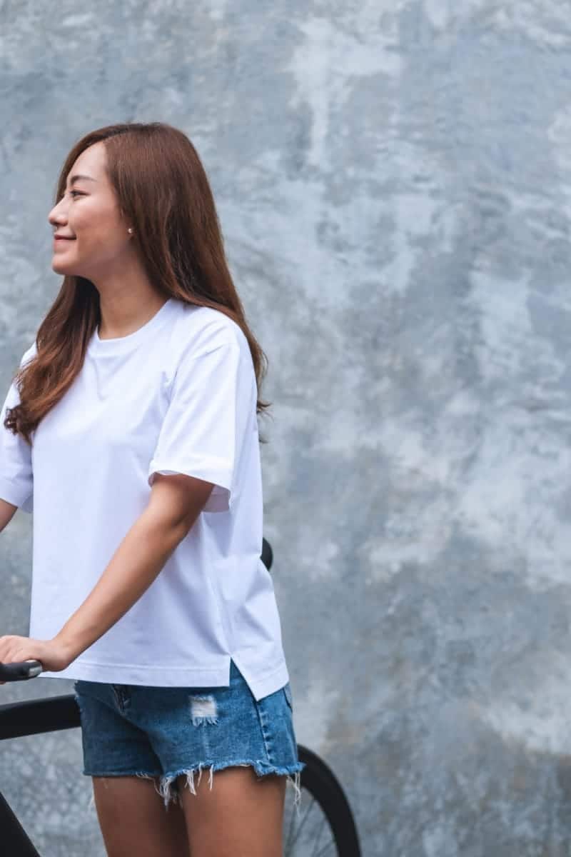 Woman wearing white tee shirt standing outside with bicycle.